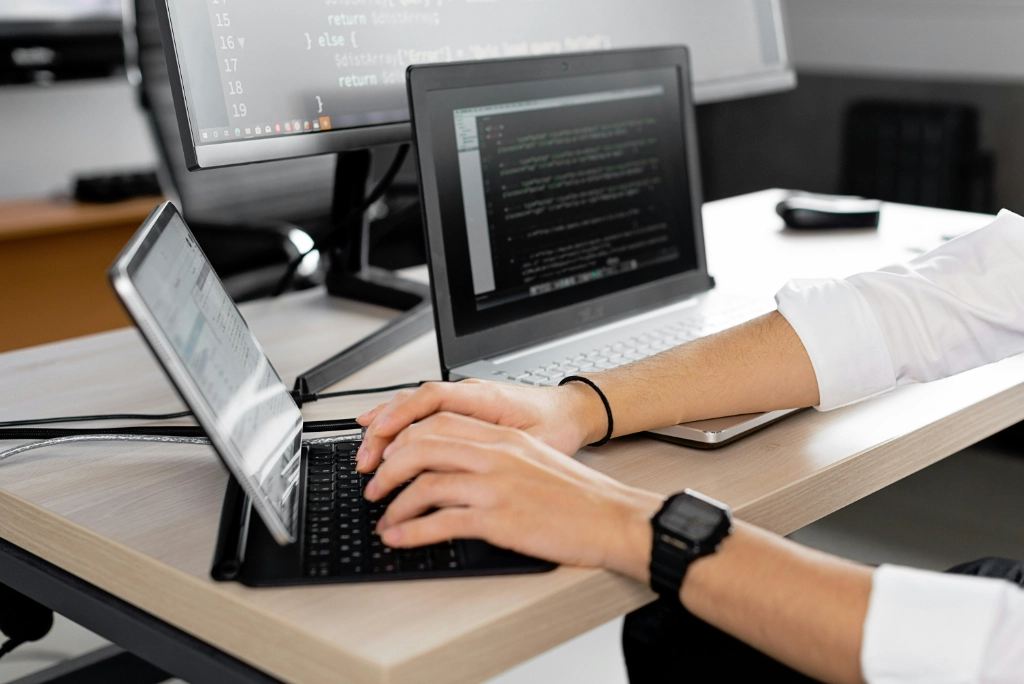 Développeur tapant sur un clavier avec plusieurs ordinateurs affichant du code à l'écran, dans un environnement de bureau moderne.